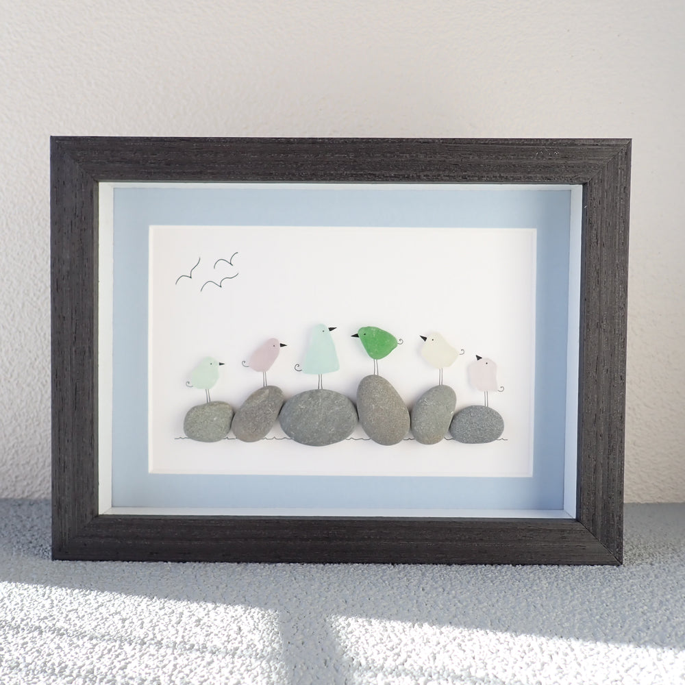 
                  
                    black shadowbox framed picture of six multicolored sea glass birds sitting on beach rocks with a light blue mat
                  
                