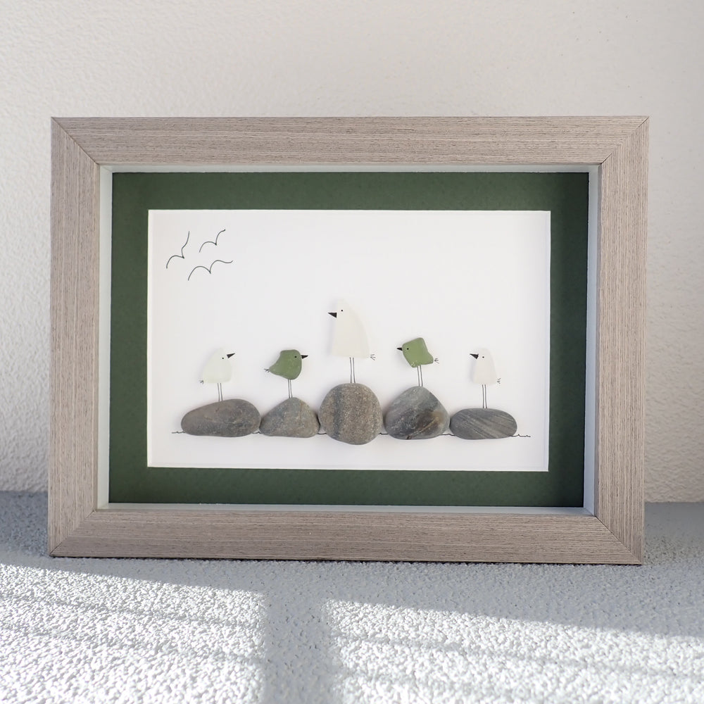 
                  
                    framed picture of six white or green colored sea glass birds sitting on beach rocks
                  
                