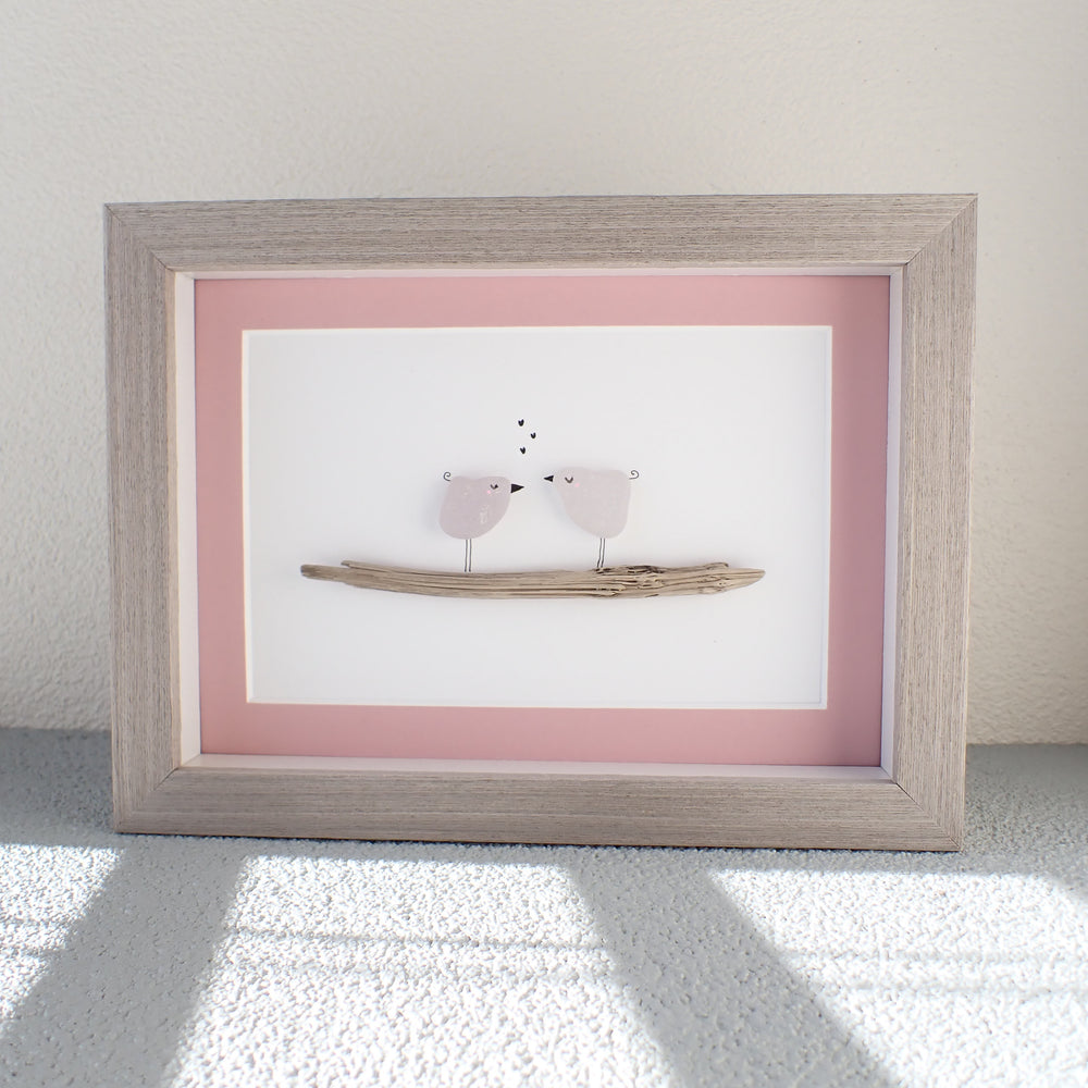 
                  
                    gray framed picture of two purple sea glass birds sitting on a piece of driftwood with a pink mat
                  
                