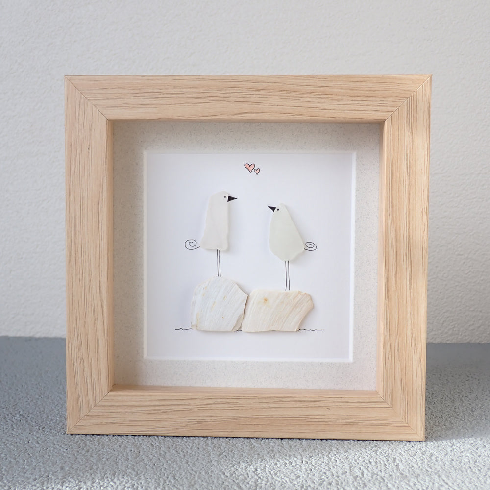 
                  
                    framed picture of two white sea glass birds standing on broken clam shells
                  
                